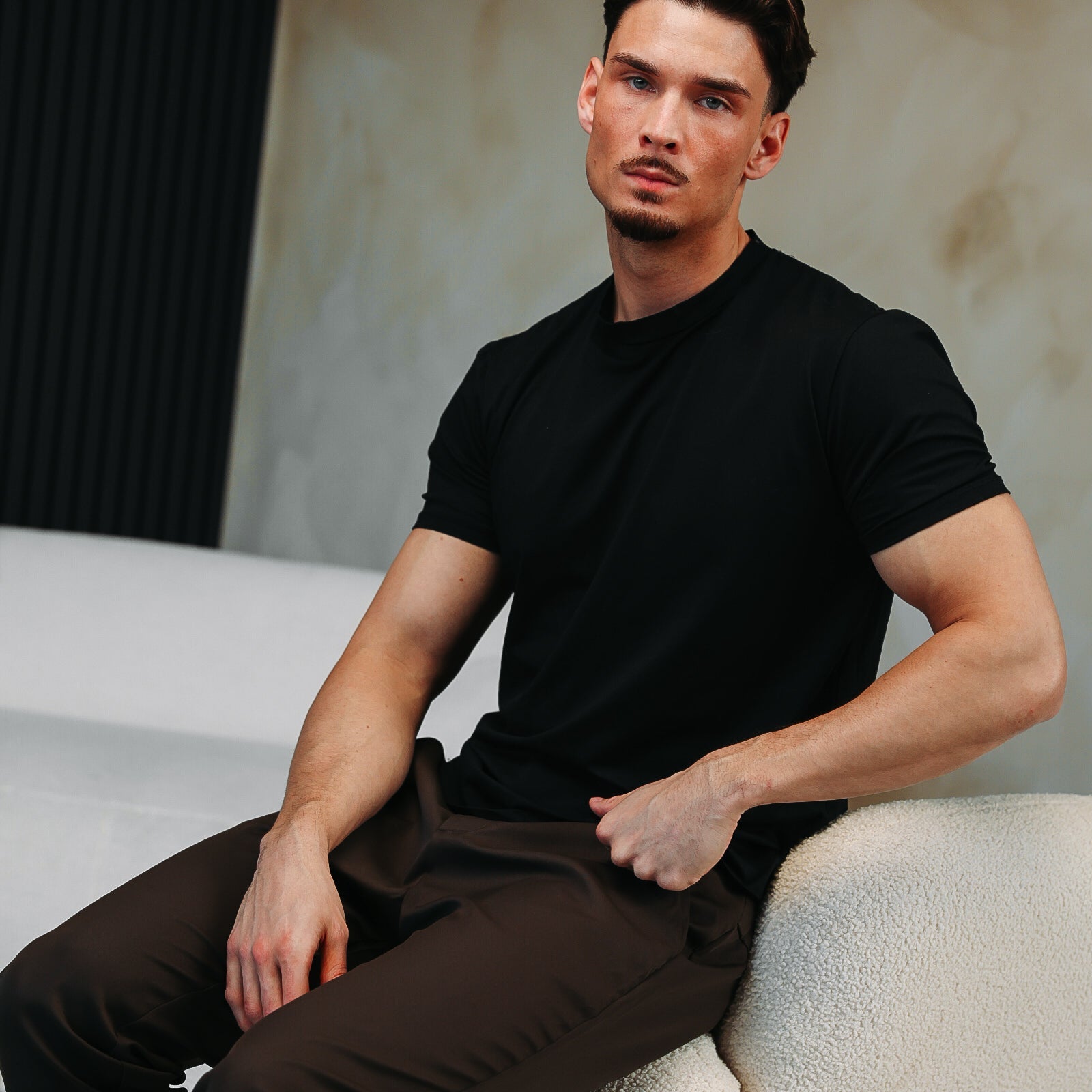 Model in het zwarte Shiny Essential Shirt, zittend op de leuning van een witte bank