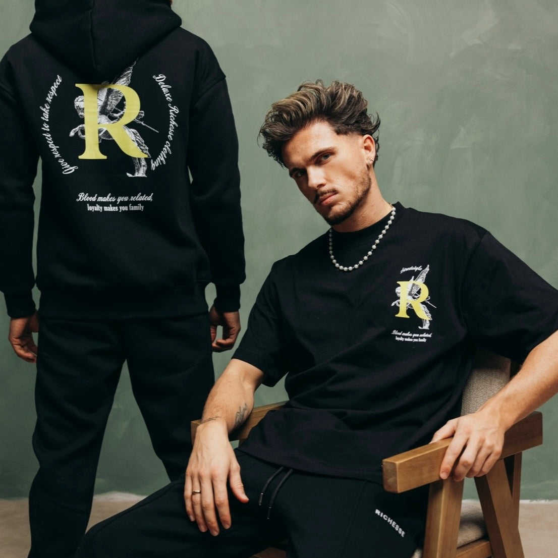 Twee modellen in zwarte Richesse-kleding met een groot R-logo, tegen een groene muur
