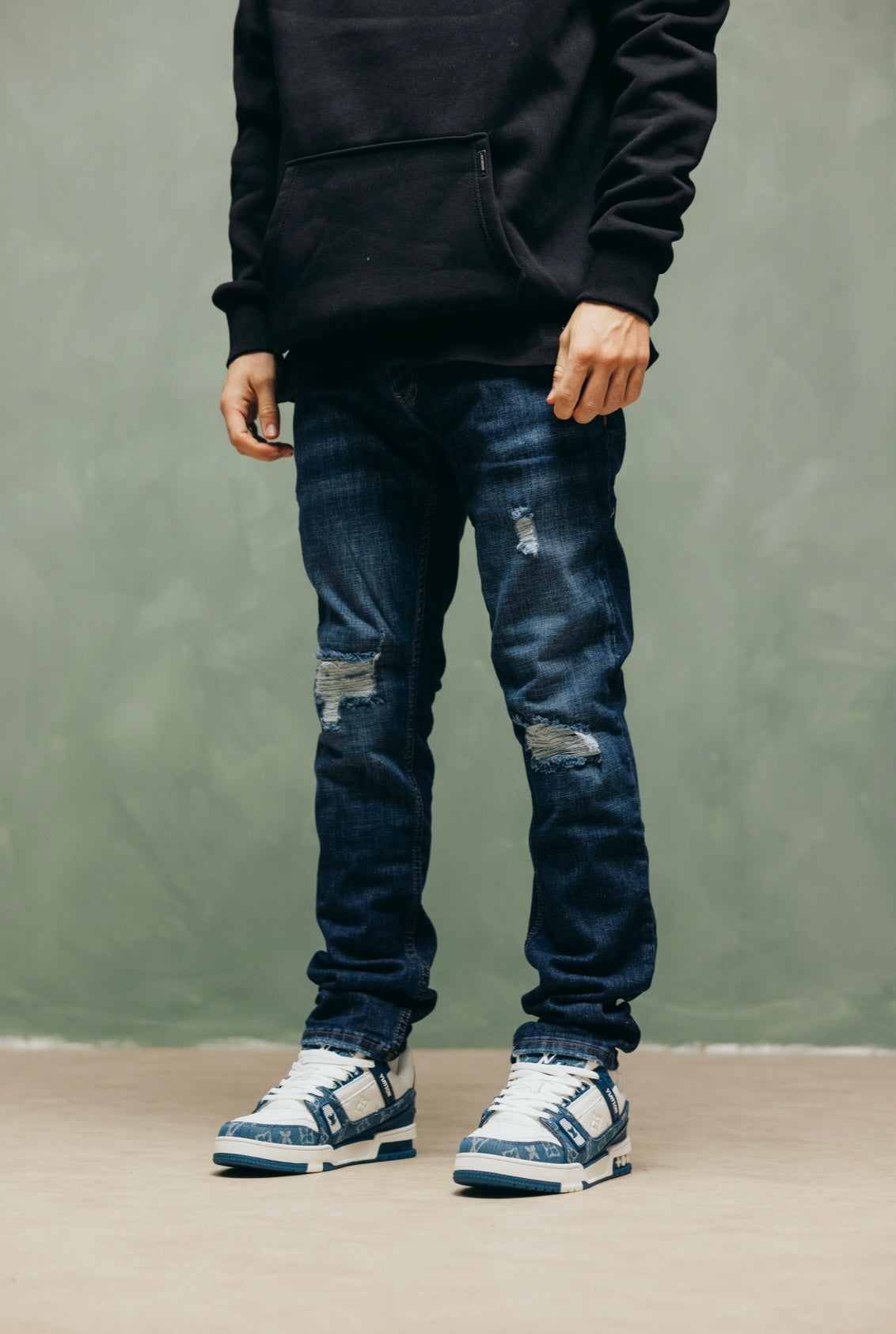 Model in de blauwe Prato Blue Jeans met sneakers, tegen een groene muur