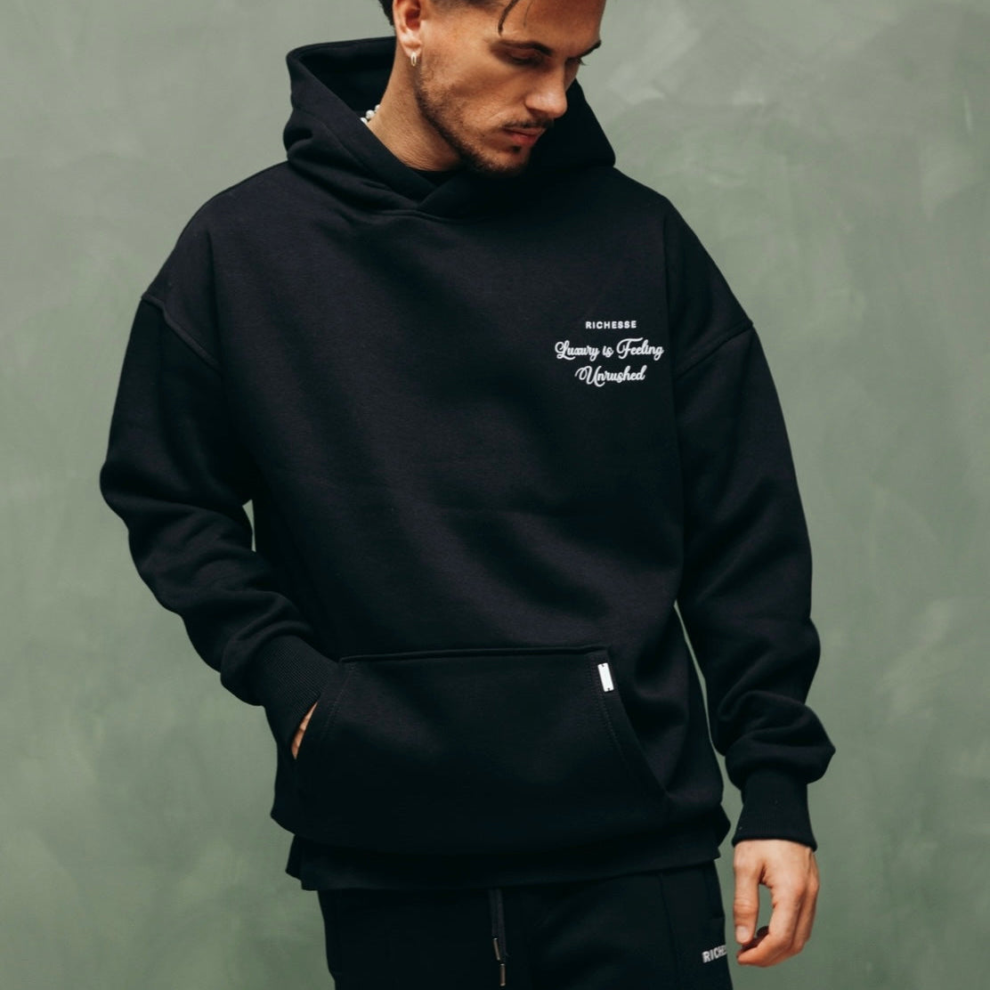 Model in de zwarte Unrushed Hoodie met print op de borst, tegen een groene muur