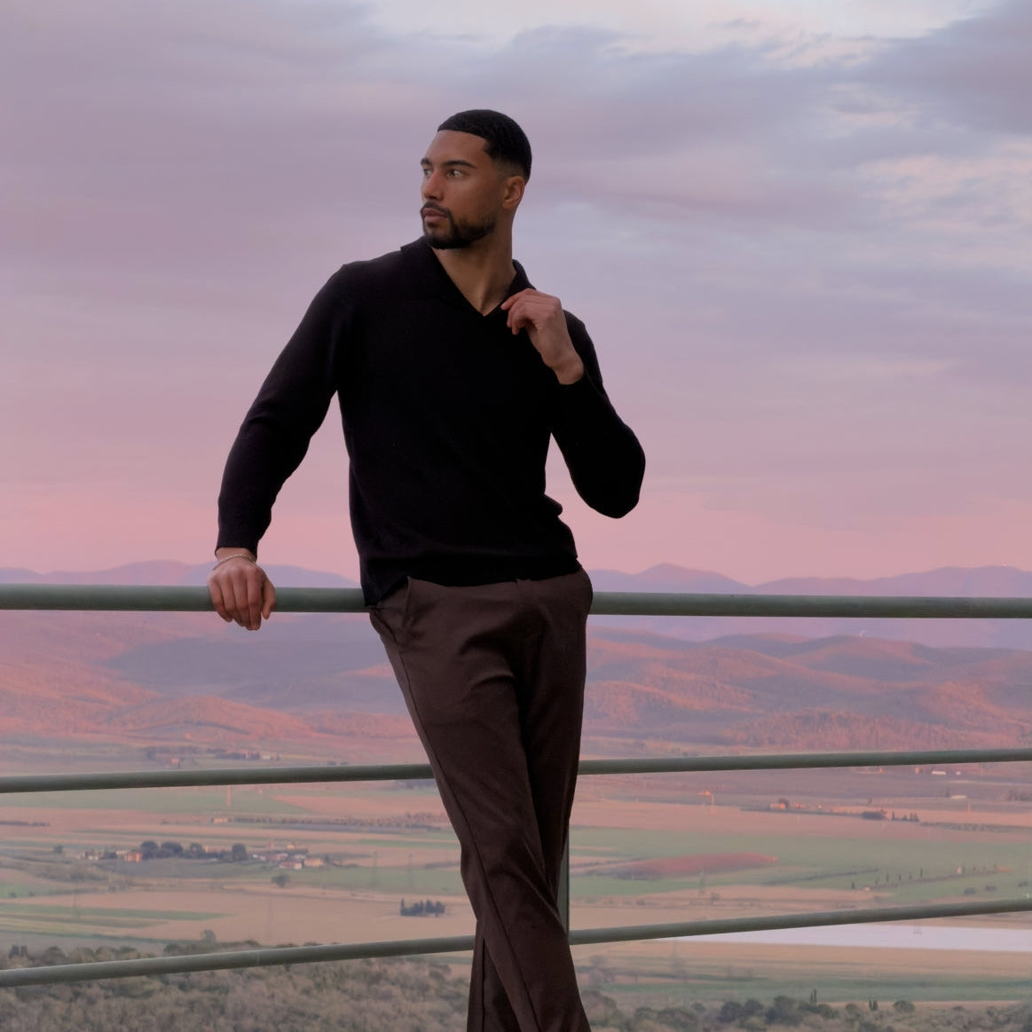 Model in de zwarte Renzo Polo met een bruine broek op een terras bij zonsondergang