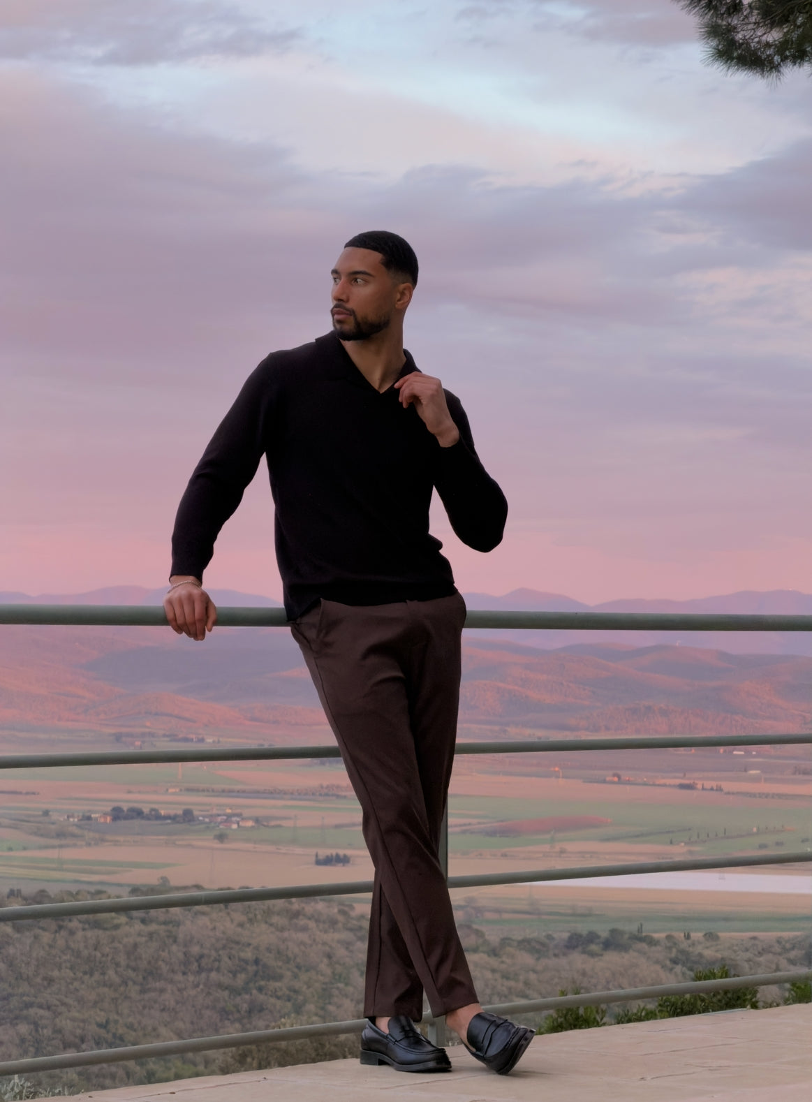 Model in de zwarte Renzo Polo met een bruine broek op een terras bij zonsondergang