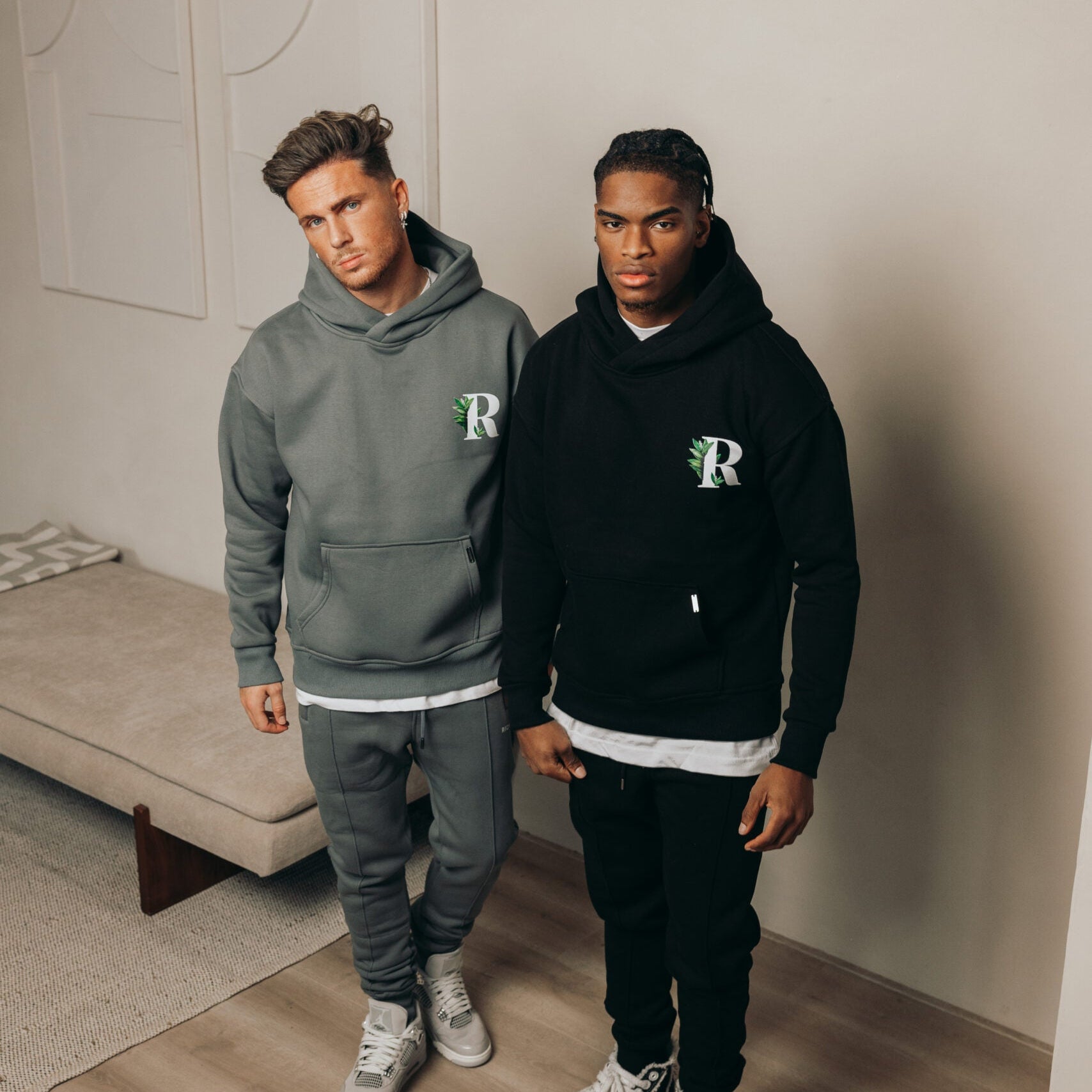 Twee modellen in een grijze en een zwarte Richesse hoodie met R-logo in een lichte kamer